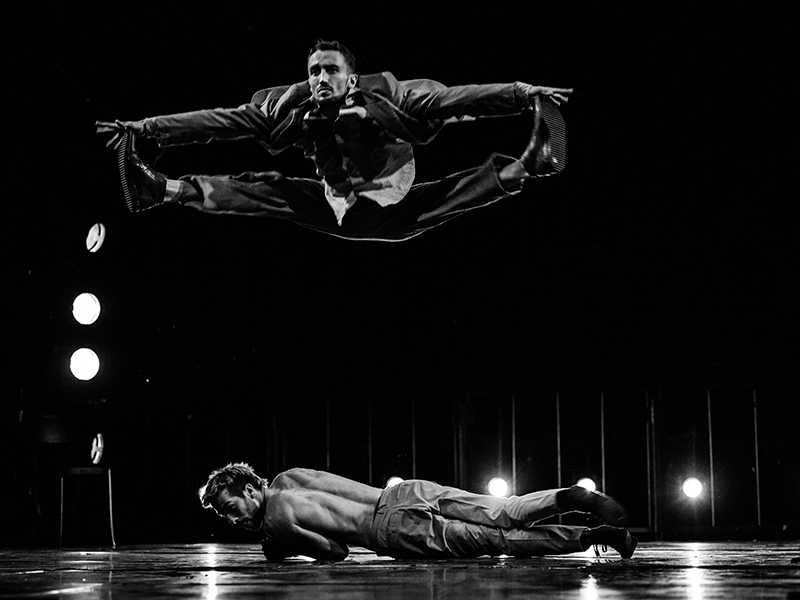 foto in bianco e nero: su un palco semibuio un uomo compie una spaccata in aria mentre un altro è sdraiato a terra sotto di lui.
