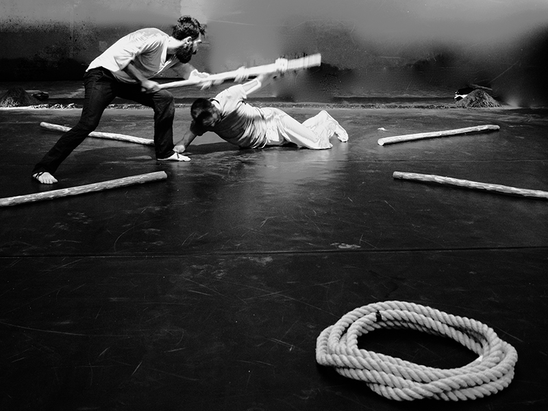 foto in bianco e nero: i due danzatori vengono immortalati mentre danzano; uno dei due è a terra. Sulla scena una corda e dei bastoni.