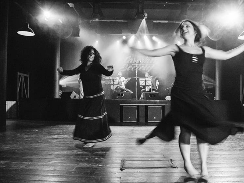 foto in bianco e nero; due danzatrici ballano con sullo sfondo i musicisti in concerto.