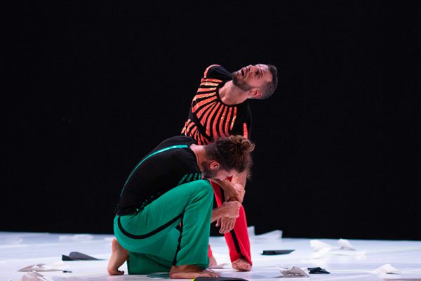 sfondo nero. Su un palco bianco i due danzatori indossano uno vestiti neri e rossi mentre l’altro vestiti neri e verdi. Ballano e sul pavimento degli aeroplanini di carta.