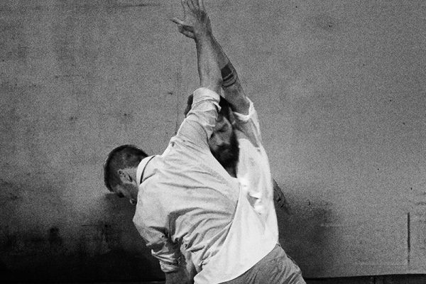 foto in bianco e nero: i due danzatori indossano camicie bianche e vengono immortalati in una posa di danza. I palmi delle loro mani si toccano.