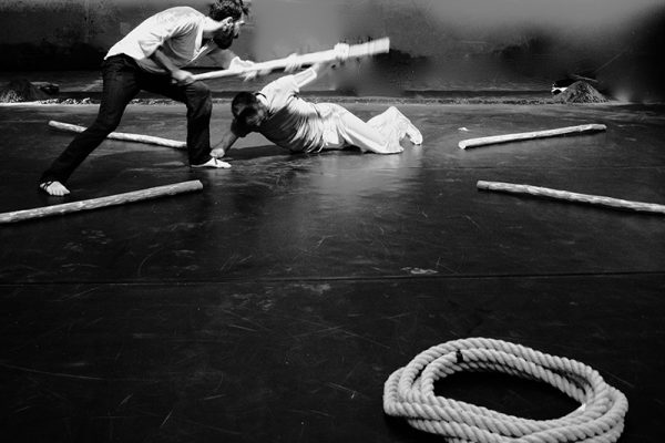 foto in bianco e nero: i due danzatori vengono immortalati mentre danzano; uno dei due è a terra. Sulla scena una corda e dei bastoni.