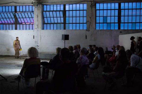 in uno spazio interno di tipologia industriale viene ripresa di scorcio parte del pubblico, in fondo il danzatore.